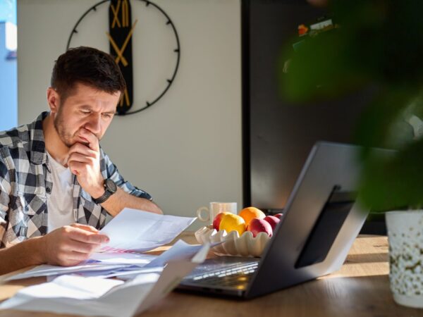 An adult man sitting at a table in his home. There are several documents on the table and a laptop open. He is holding one document in his hand and looking at it with a concerned, confused expression.