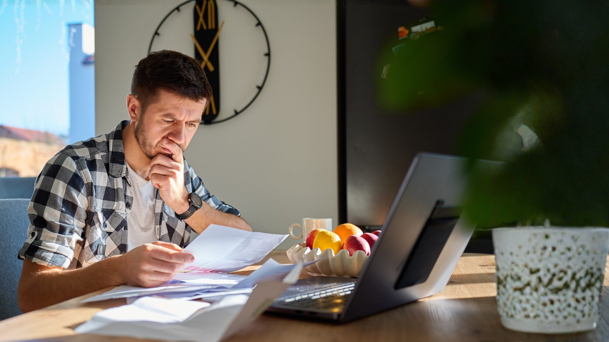 An adult man sitting at a table in his home. There are several documents on the table and a laptop open. He is holding one document in his hand and looking at it with a concerned, confused expression.