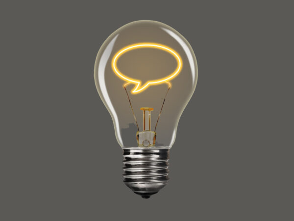 A photo of a lightbulb on the grey background. Instead of the regular lightbulb filament, the filament is in the shape of a speech bubble. It is glowing yellow.